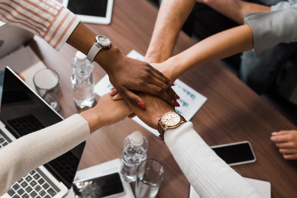 částečný pohled na multikulturní podnikatele držící se za ruce nad stolem s pomůckami, pohled shora - Fotografie, Obrázek