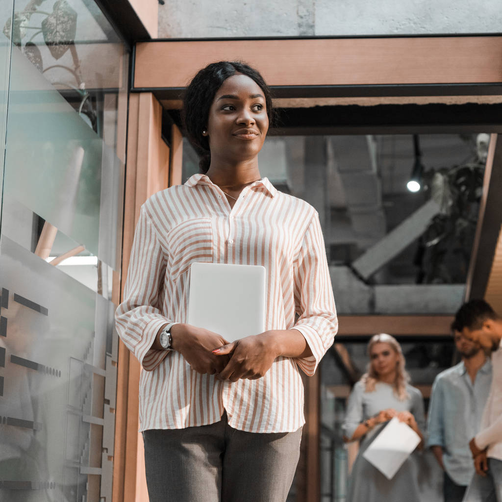 счастливая африканская американская бизнесвумен смотрит в сторону и держит цифровой планшет в то время как мультикультурные коллеги стоят на заднем плане
 - Фото, изображение