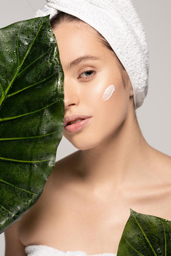 young woman with moisturizing cream on face and towel on head posing with green leaves, isolated on grey - Photo, Image