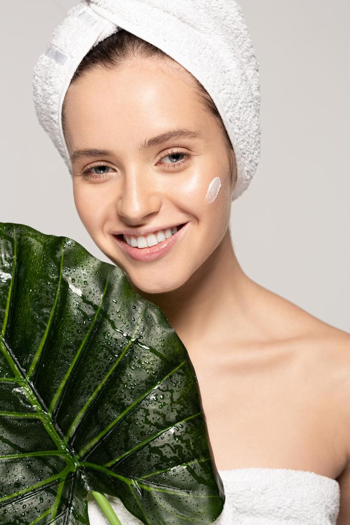 χαρούμενο κορίτσι με ενυδατική κρέμα στο πρόσωπο και πετσέτα στο κεφάλι ποζάροντας με πράσινο φύλλο, απομονωμένο σε γκρι - Φωτογραφία, εικόνα