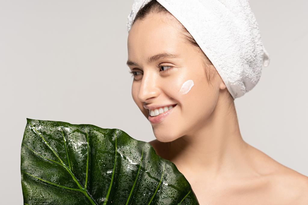 fröhliches Mädchen mit kosmetischer Creme im Gesicht und Handtuch auf dem Kopf posiert mit grünem Blatt, isoliert auf grau  - Foto, Bild