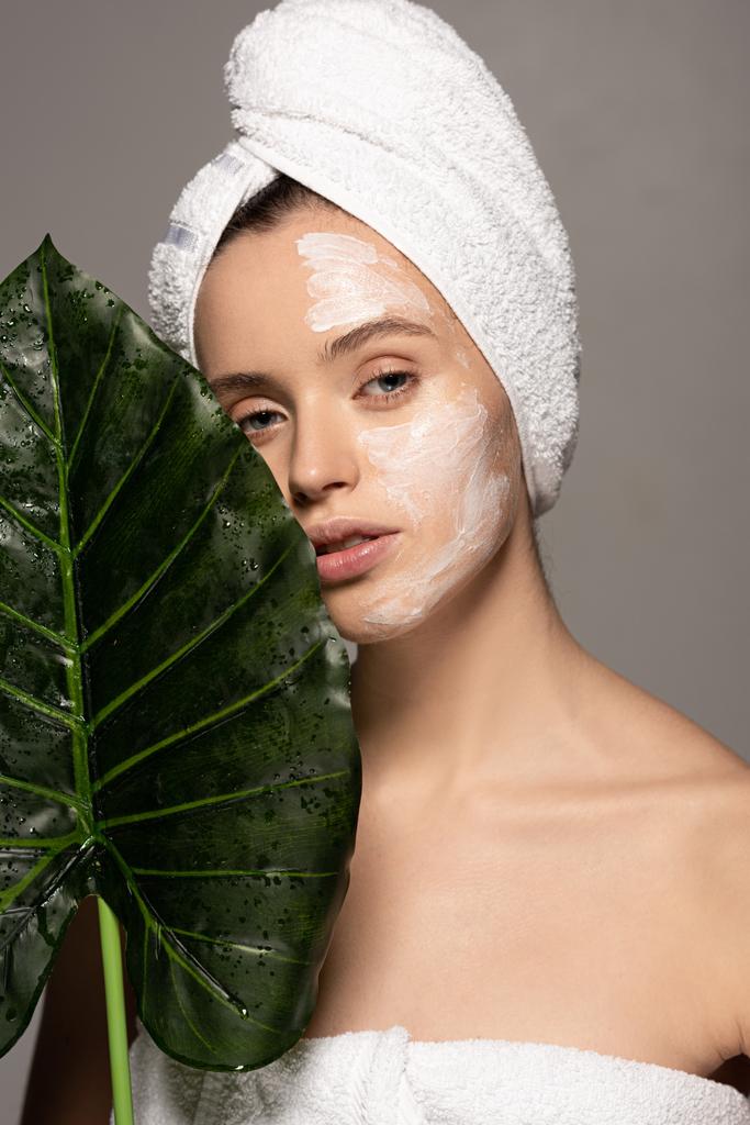 atrakcyjna dziewczyna z maską kosmetyczną na twarzy i ręcznikiem na głowie pozowanie z zielonym liściem, odizolowana na szaro - Zdjęcie, obraz