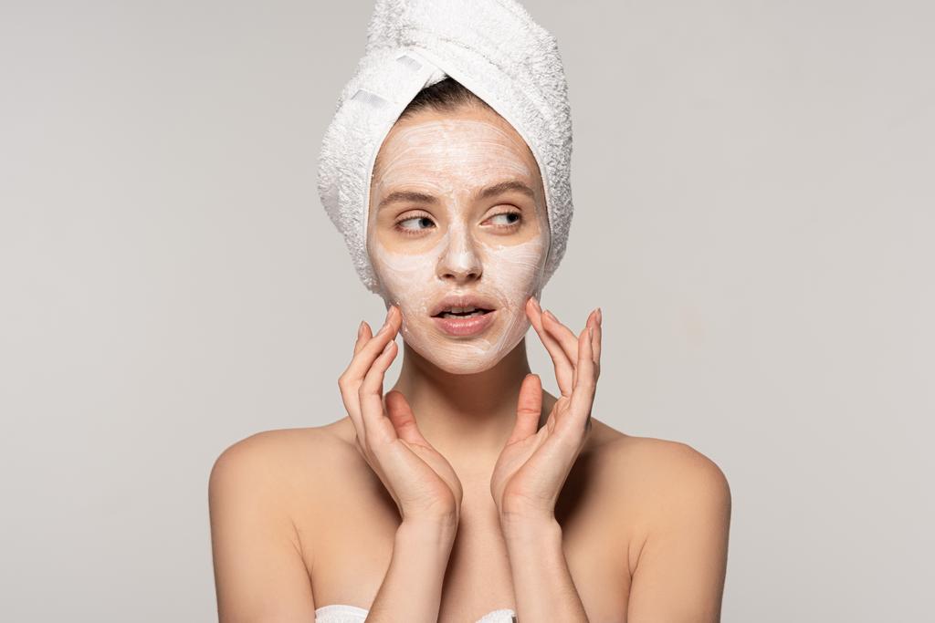 atraktivní zamyšlená dívka s kosmetickou maskou na obličeji a ručníkem na hlavě, izolovaná na šedé  - Fotografie, Obrázek