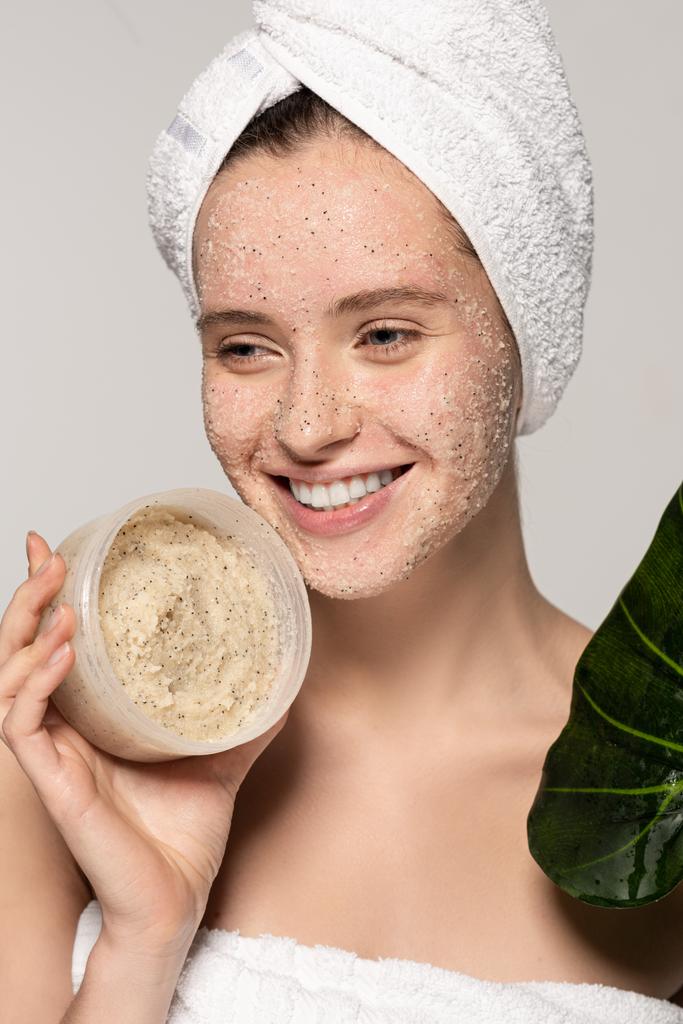 radosna kobieta z ręcznikiem na głowie trzymająca zielony liść i plastikowy pojemnik z peelingiem kokosowym, odizolowana na szaro - Zdjęcie, obraz