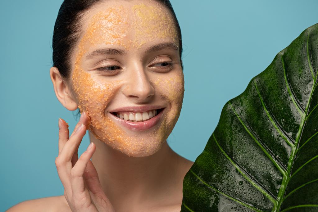 smiling girl with sugar scrub on face holding leaf, isolated on blue - Photo, Image
