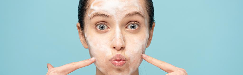 πανοραμική λήψη αστεία γυναίκα με αφρό καθαρισμού στο πρόσωπο δείχνει στα μάγουλα, απομονώνονται σε μπλε - Φωτογραφία, εικόνα