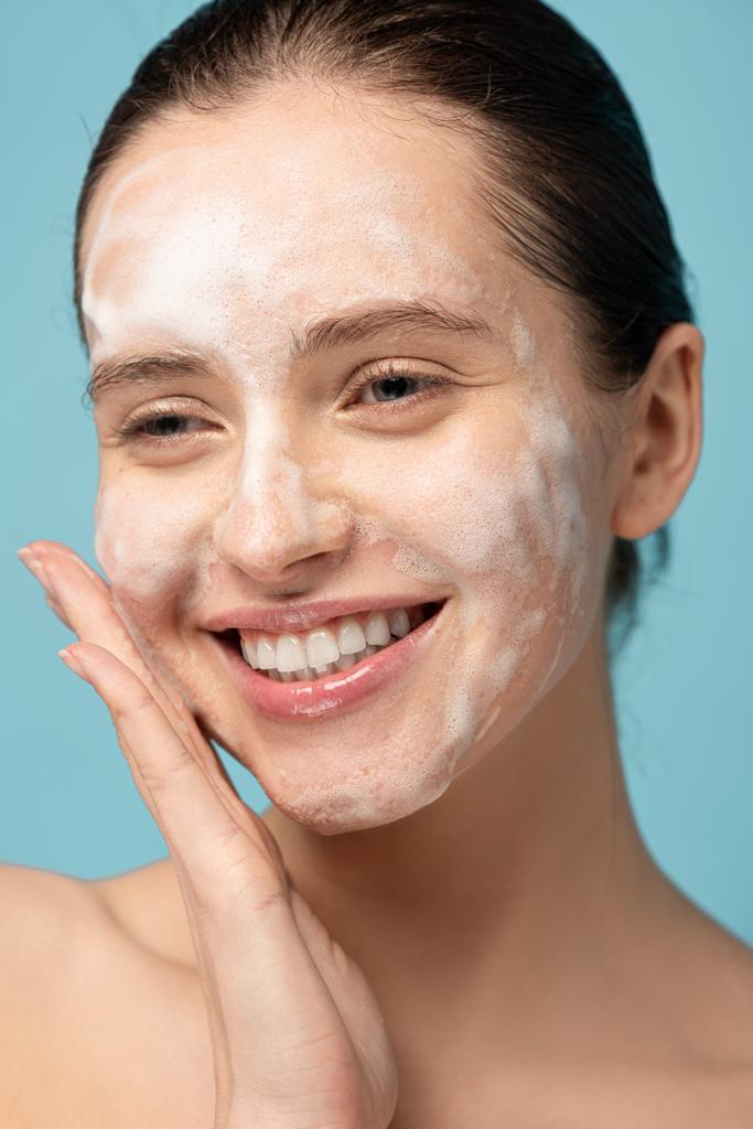 attractive happy girl applying cleansing foam on face, isolated on blue - Photo, Image