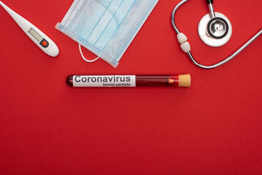 Vista superior de la máscara médica con termómetro, estetoscopio y letras de coronavirus en el tubo de ensayo con sangre sobre fondo rojo
 - Foto, Imagen
