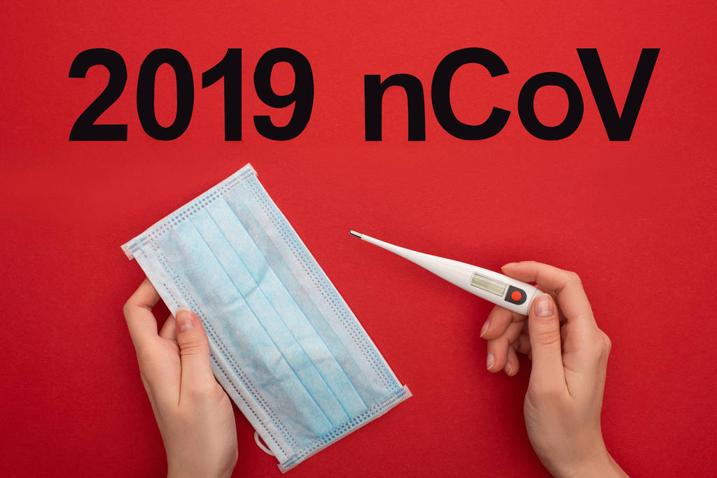 Top view of woman holding medical mask with thermometer near 2019 ncov lettering on red surface - Photo, Image
