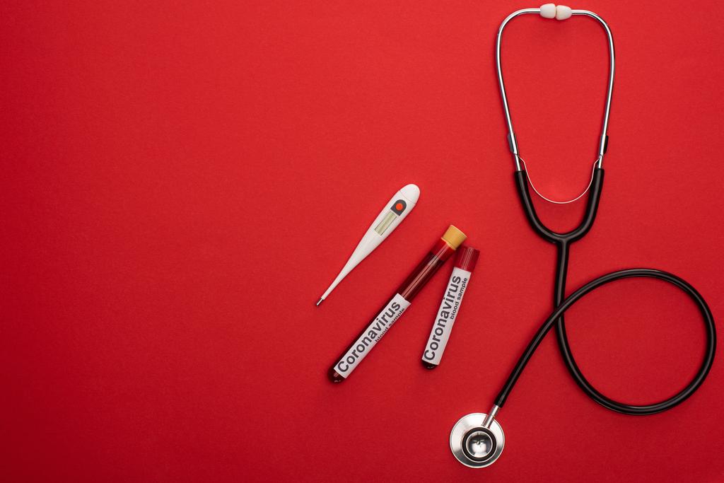 Horní pohled na stetoskop, teploměr a zkumavky s krevními vzorky a koronavirem na červeném pozadí - Fotografie, Obrázek