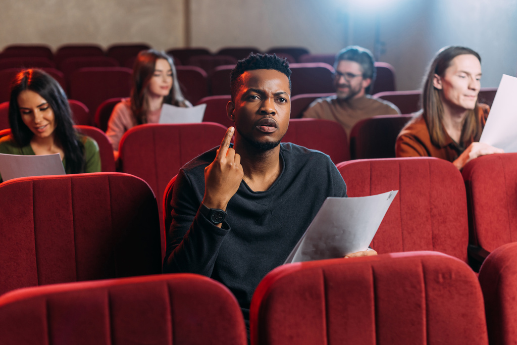 Afrikanischer amerikanischer Schauspieler probt und zeigt nach oben, während er mit Schauspielern auf roten Stühlen sitzt - Foto, Bild