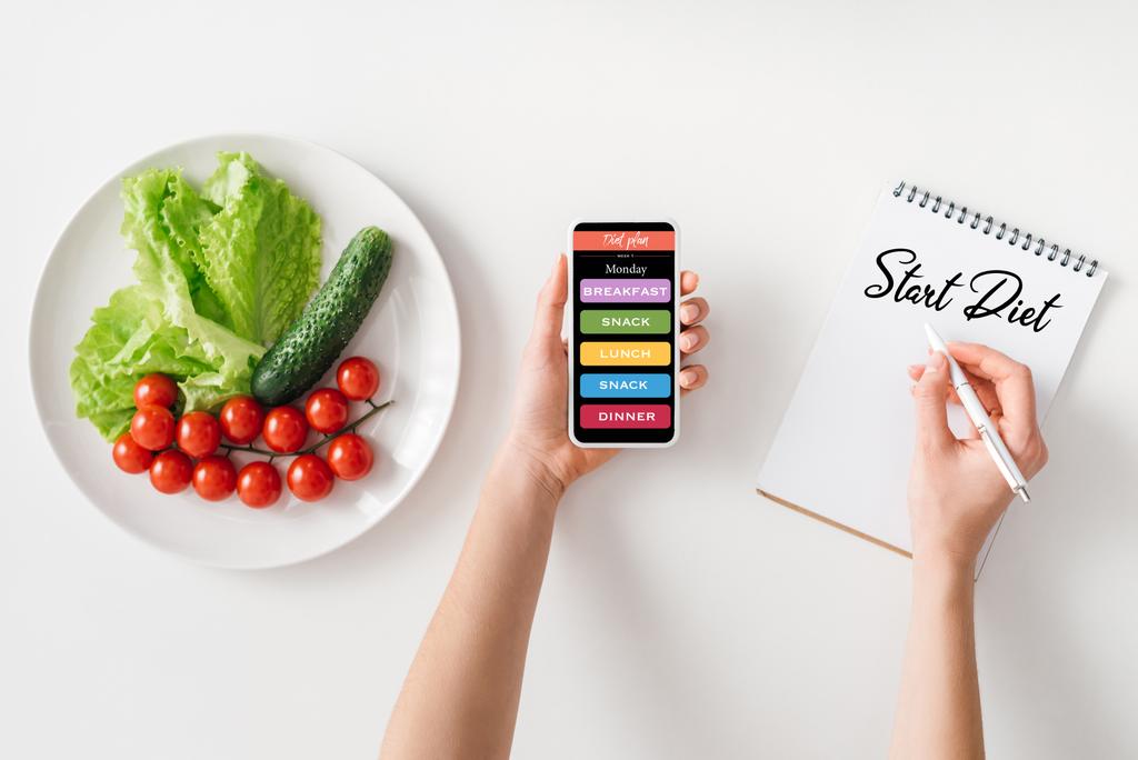 Bovenaanzicht van vrouw met smartphone en schrijven in notebook met start dieet belettering in de buurt van rijpe groenten op plaat op witte achtergrond - Foto, afbeelding