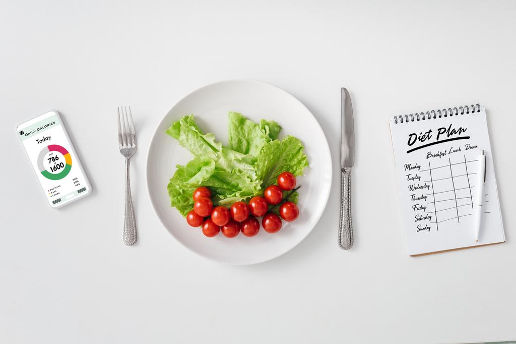 皿の上の新鮮な野菜のトップビュー,白い背景にダイエット計画とカロリーカウントアプリやノートブックを持つスマートフォン. - 写真・画像
