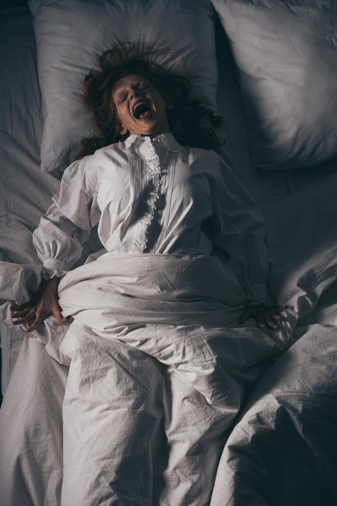 Вид сверху на демоническую злую девушку в ночном халате, кричащую, лежа в постели
 - Фото, изображение