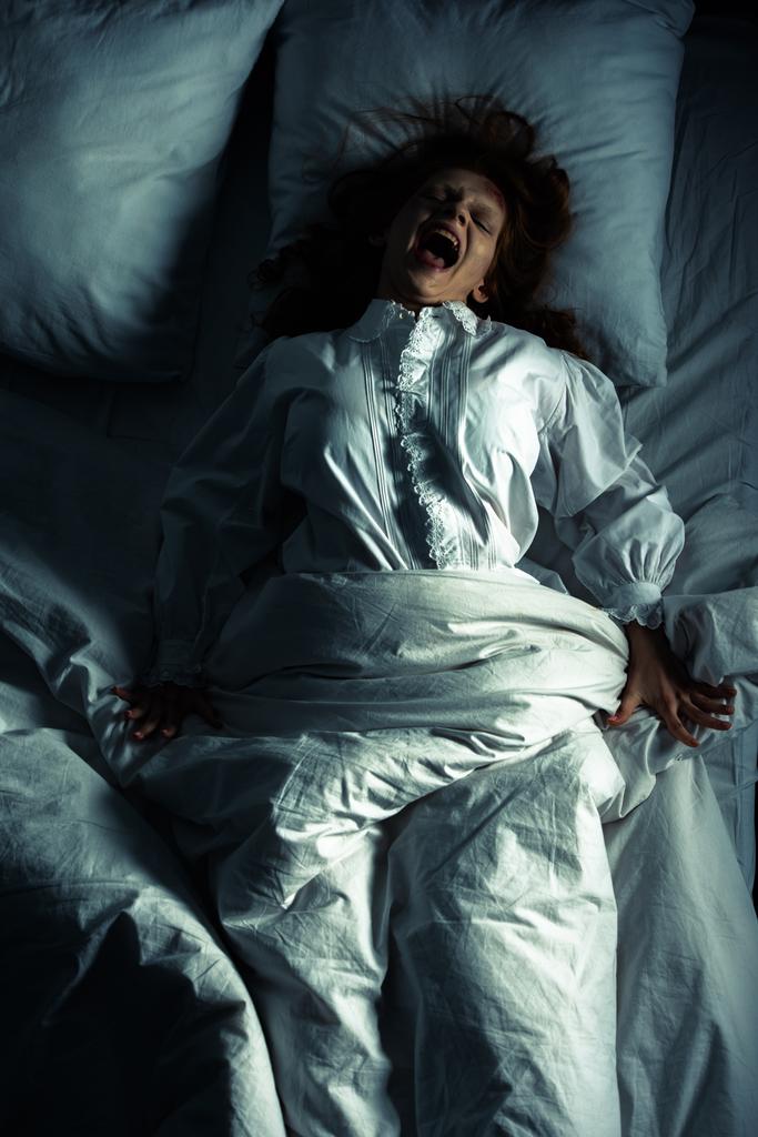 κορυφαία άποψη του θηλυκό δαίμονα σε νυχτικό ουρλιάζοντας, ενώ βρίσκεται στο κρεβάτι - Φωτογραφία, εικόνα