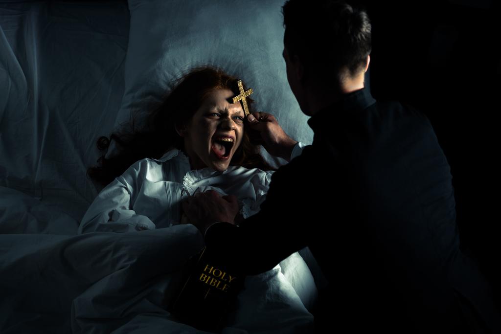 聖書を持ったエクソシストとベッドの中で悪魔の叫びの少女の上に立って - 写真・画像