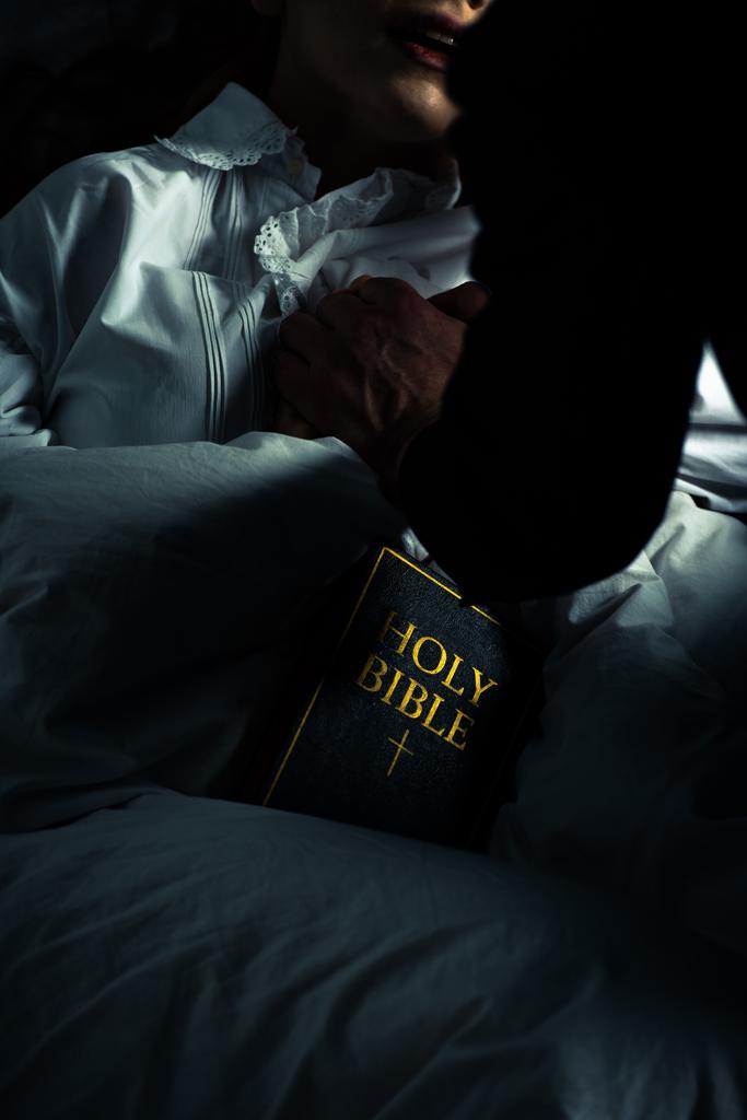περικοπή άποψη του εξορκιστή με Αγία Γραφή κρατώντας τα χέρια με εμμονή κορίτσι στο κρεβάτι  - Φωτογραφία, εικόνα