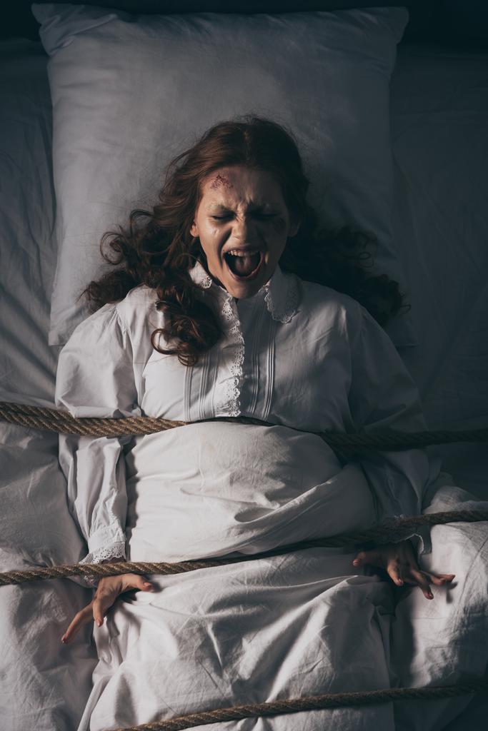 Dämonisch besessenes schreiendes Mädchen im Nachthemd, im Bett mit Seil gefesselt - Foto, Bild