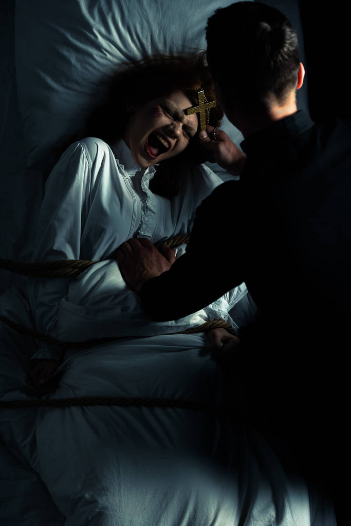 egzorcysta trzyma krzyż nad kobiecym demonem związany liną na łóżku  - Zdjęcie, obraz