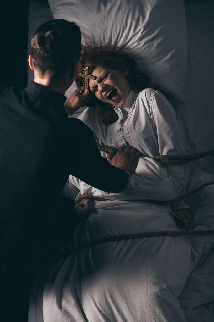 exorcista drží kříž přes démonickou posedlou dívku svázanou provazem na posteli  - Fotografie, Obrázek
