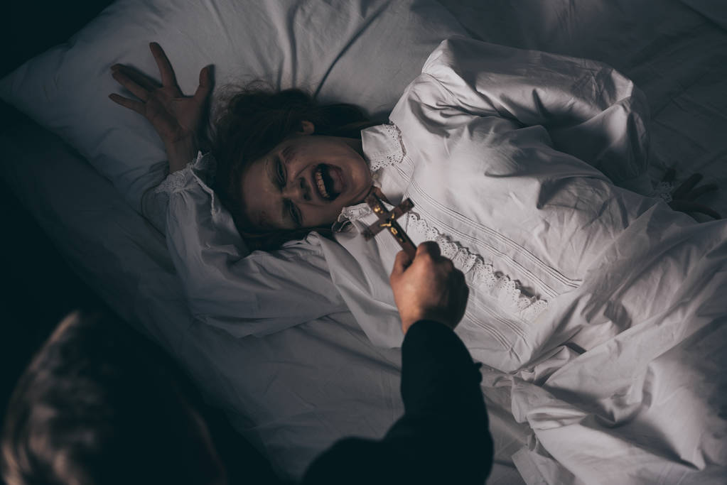 exorcista sosteniendo cruz sobre demoníaca gritando chica en la cama
 - Foto, Imagen