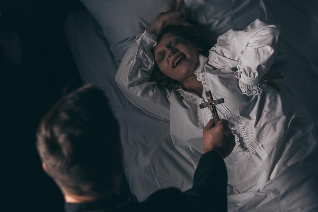 ördögűző tartja kereszt felett démoni sikoltozó lány az ágyban - Fotó, kép
