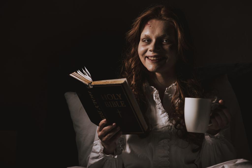 demonisch lachend meisje in nachtjapon met bijbel en beker op bed - Foto, afbeelding