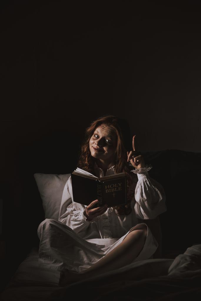 δαιμονική κοπέλα με νυχτικό που διαβάζει τη Βίβλο και δείχνει πάνω στο κρεβάτι. - Φωτογραφία, εικόνα