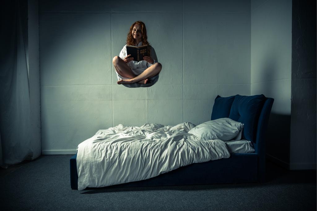 assustador sorrindo menina em camisola levitando sobre a cama enquanto lendo bíblia
 - Foto, Imagem