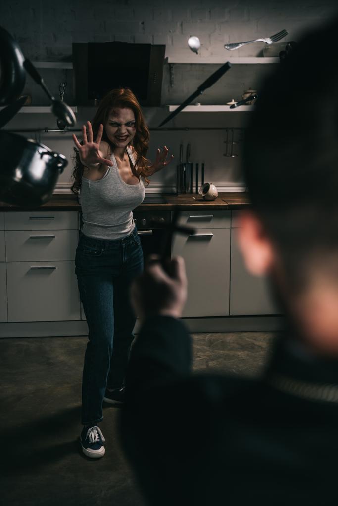 selective focus of creepy demoniacal girl with levitating cookware and exorcist with cross in kitchen - Photo, Image