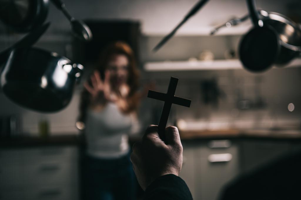selektiver Fokus weiblicher Dämonen mit schwebendem Kochgeschirr und Exorzist mit Kreuz in der Küche - Foto, Bild