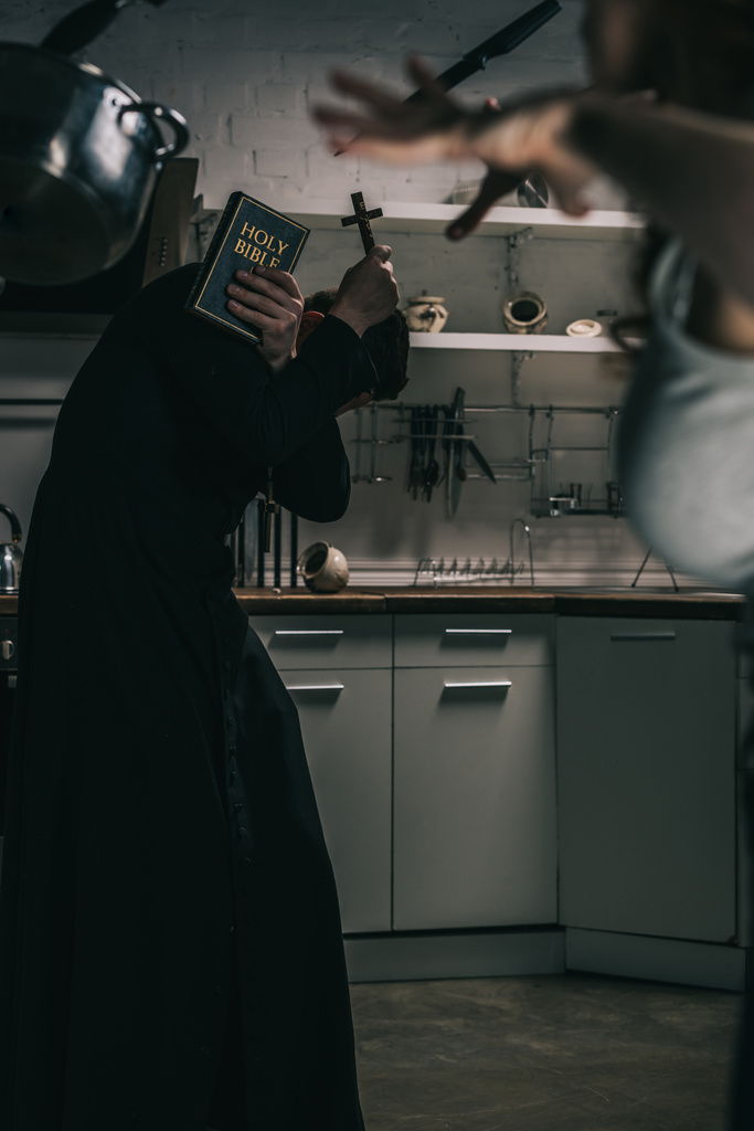 selektywne skupienie złego demona z lewitującymi naczyniami i egzorcystą z krzyżem i biblią w kuchni - Zdjęcie, obraz