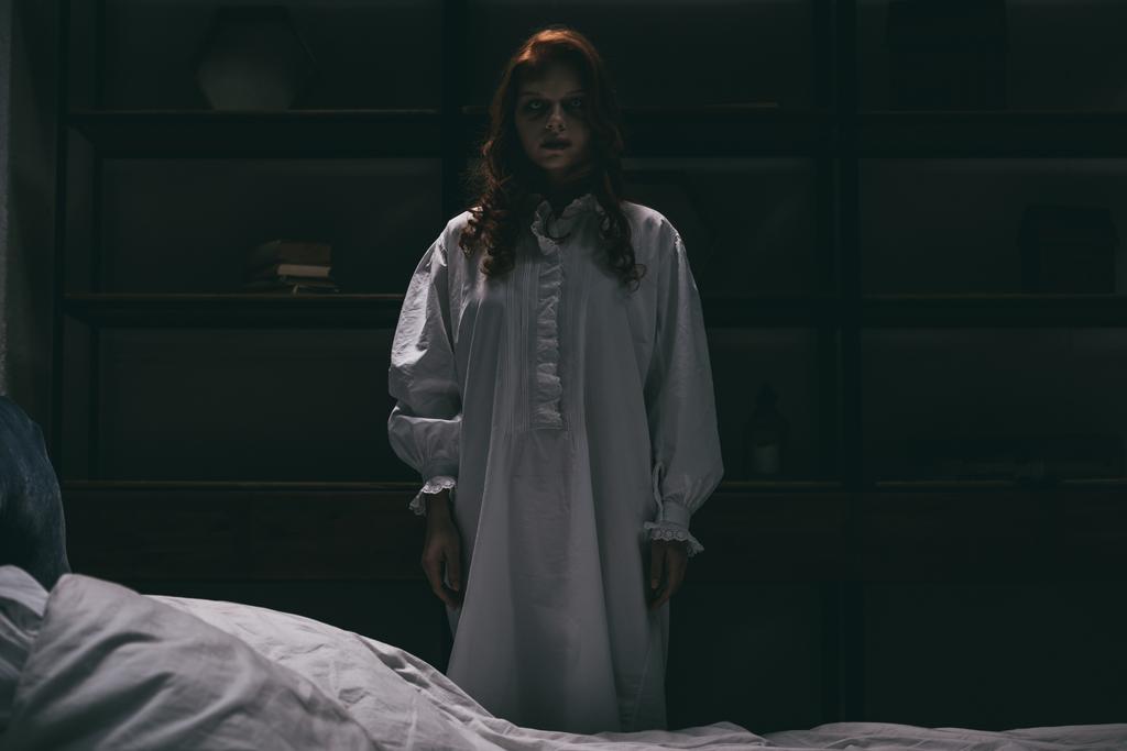 geobsedeerde vrouw in nachtjapon staan in de buurt van bed in donkere kamer - Foto, afbeelding