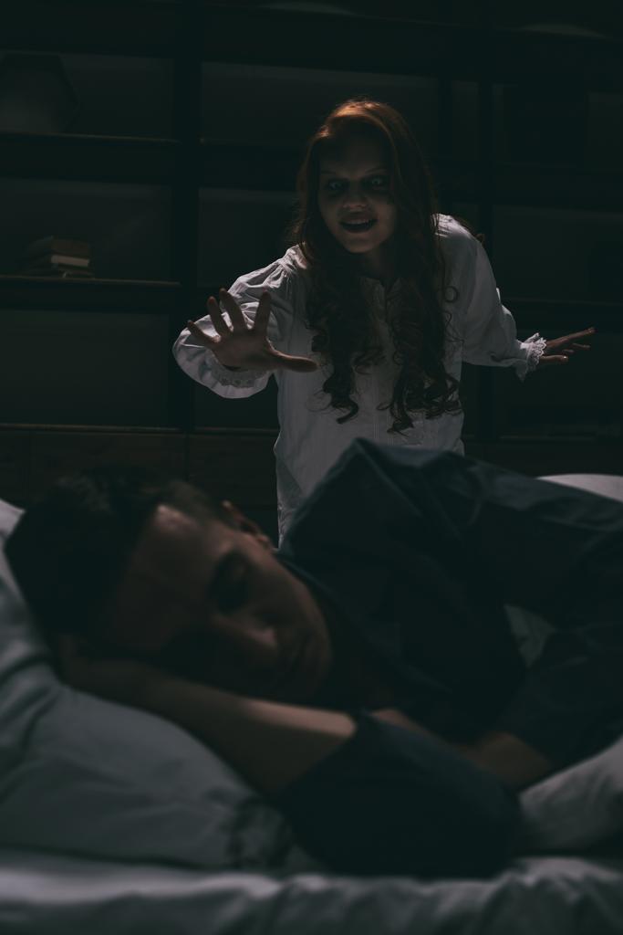 δαιμονική χαμογελαστή γυναίκα στέκεται πάνω από τον κοιμισμένο άντρα στην κρεβατοκάμαρα  - Φωτογραφία, εικόνα
