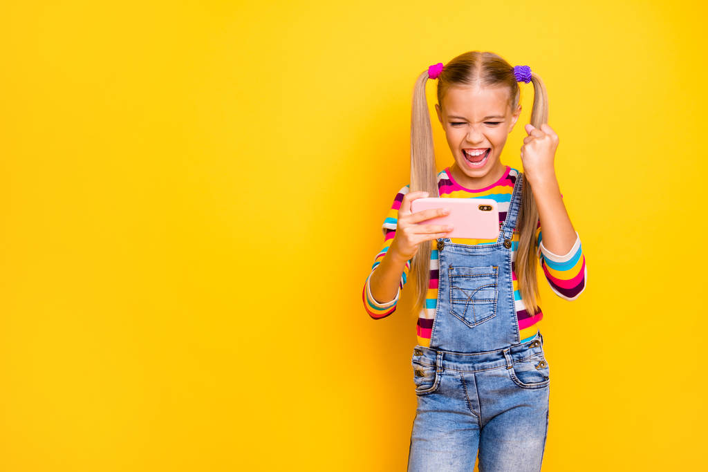 Портрет екстатичної збудженої дівчинки використовує смартфон грати онлайн відеоігри підняти кулаки кричать так, носити смугастий светр джинсові джинси ізольовані на жовтому яскравому кольоровому фоні
 - Фото, зображення