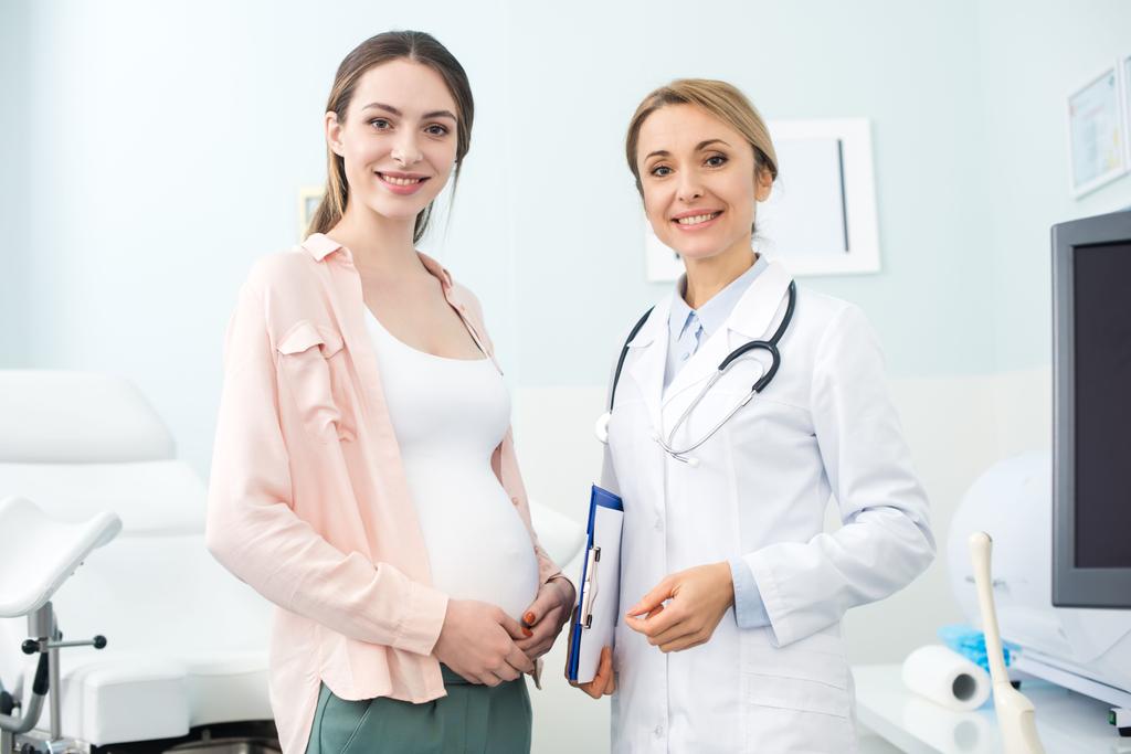 Junge Schwangere steht mit lächelndem Gynäkologen in Klinik - Foto, Bild