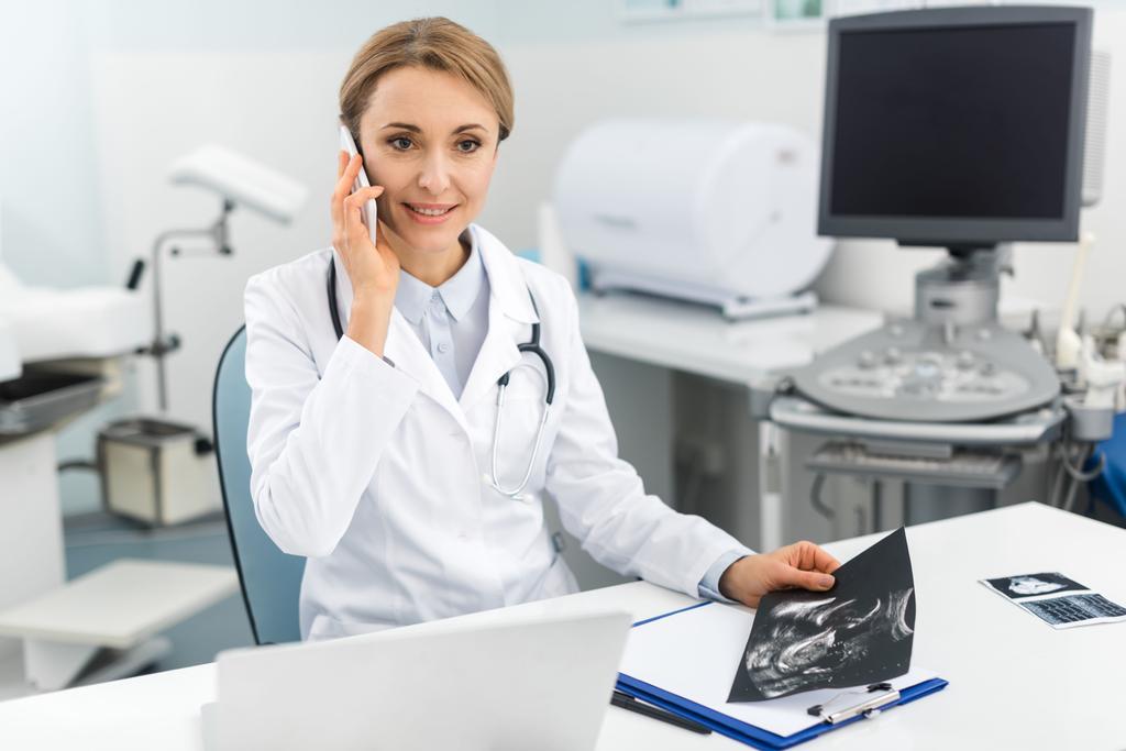 médico sonriente profesional sosteniendo ultrasonido escaneo y hablando en el teléfono inteligente en la clínica
  - Foto, Imagen