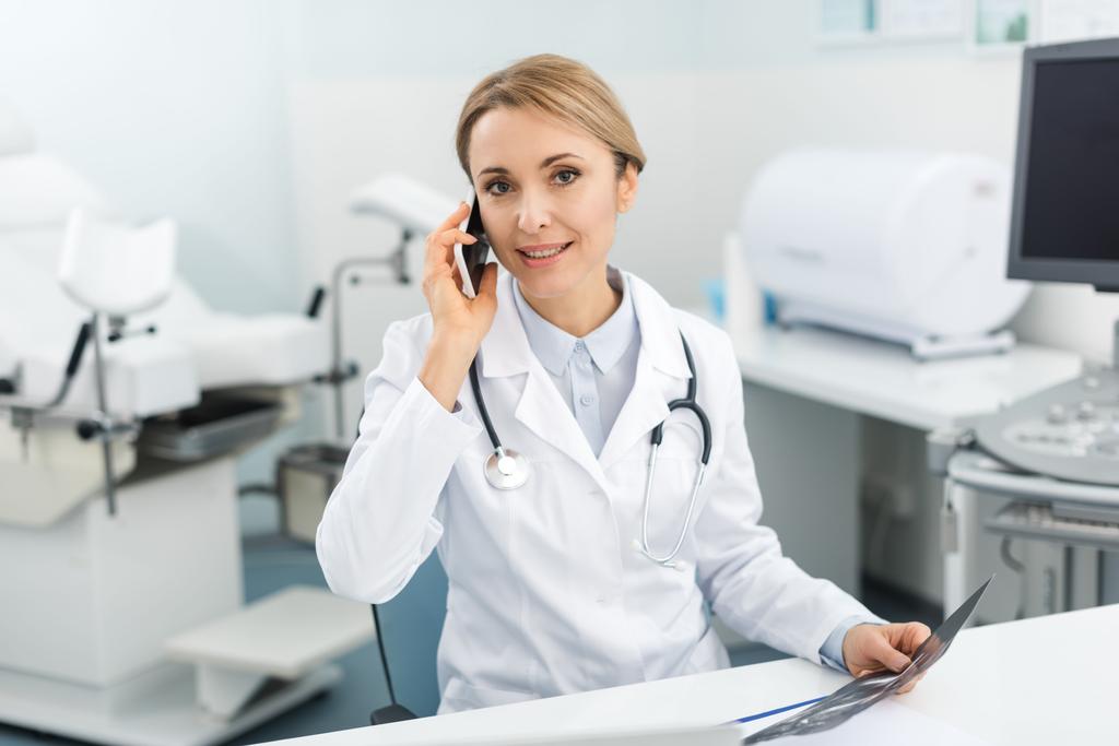 doctora profesional sosteniendo ultrasonido escaneo y hablando en teléfono inteligente en la clínica
  - Foto, Imagen