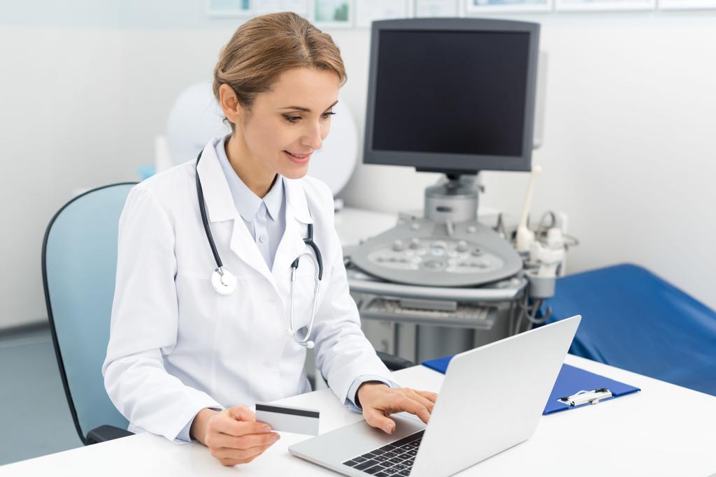 usmívající se lékař pomocí notebooku a kreditní karty na klinice s ultrazvukovým skenerem - Fotografie, Obrázek