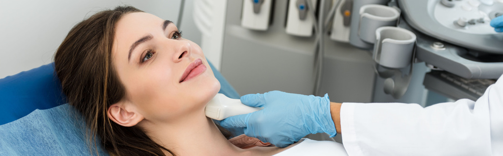 panoramisch schot van professionele arts onderzoeken schildklier van vrouwelijke patiënt met echografie scan in kliniek - Foto, afbeelding