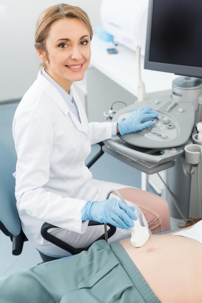 Gülümseyen doktor, klinikte ultrason taramasıyla hamile kadının karnını inceliyor. - Fotoğraf, Görsel