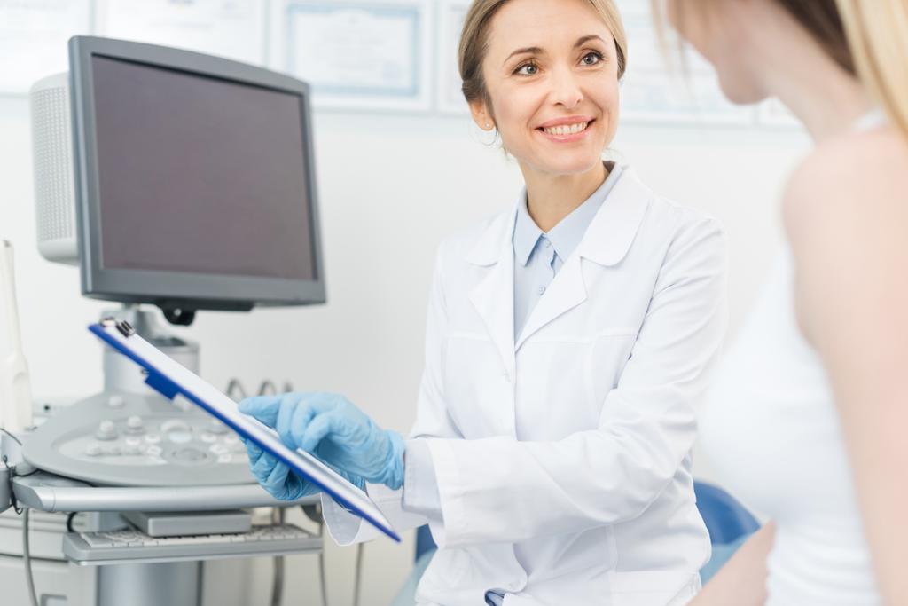 glimlachende arts toont diagnose aan patiënt in kliniek in de buurt van echografie scanner - Foto, afbeelding