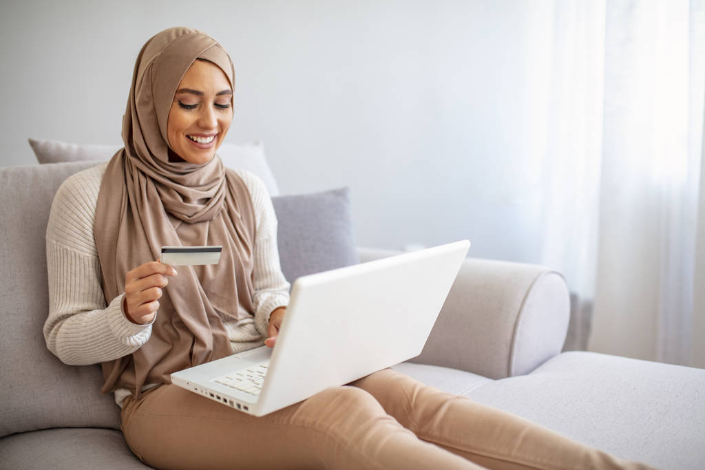 Porträt einer schönen jungen muslimischen Frau, die auf ihrem Laptop im Internet surft, während sie im Wohnzimmer eine Kreditkarte hält. Wunderschöne junge Frau mit Laptop online einkaufen. - Foto, Bild