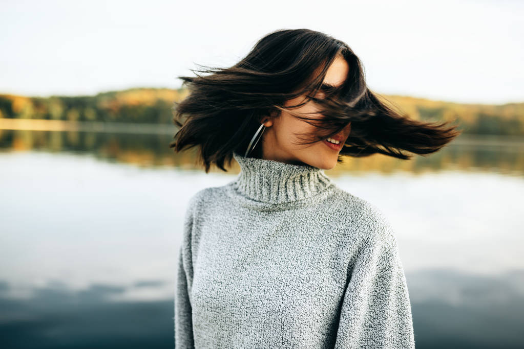 Obraz szczęśliwej młodej brunetki z zamkniętymi oczami, noszącej szary sweter, pozującej na tle natury. Ładna dziewczyna pozuje na jezioro w parku. - Zdjęcie, obraz