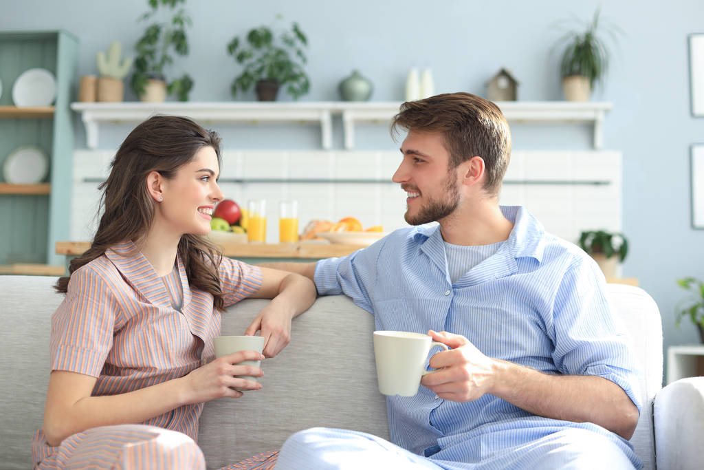 Schönes junges Paar im Pyjama schaut einander an und lächelt auf einem Sofa im Wohnzimmer. - Foto, Bild