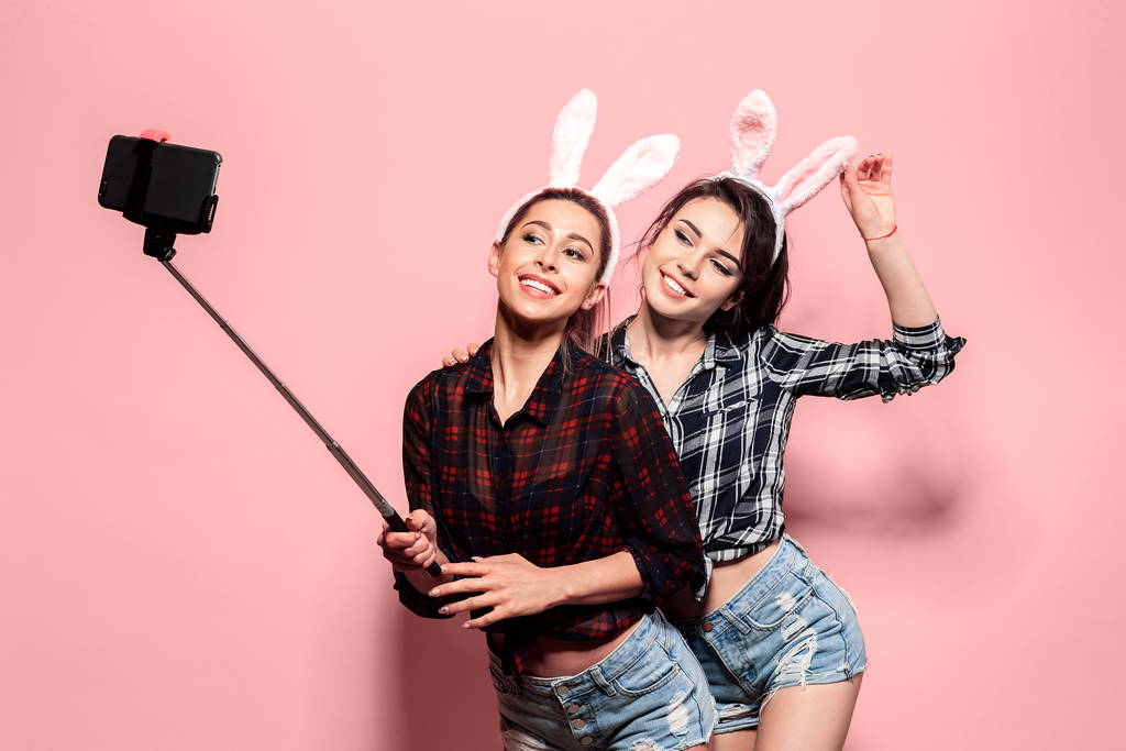 Zwei junge sexy kaukasische Frau trägt niedliche Osterhasenohren macht Selfie mit Stock über rosa isolierten Hintergrund - Foto, Bild