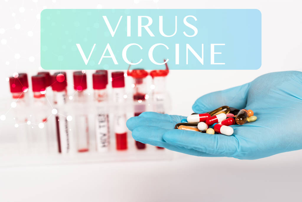 обрезанный взгляд ученого в латексной перчатке с таблетками рядом с пробирками и вирусной вакциной с надписью на белом
  - Фото, изображение