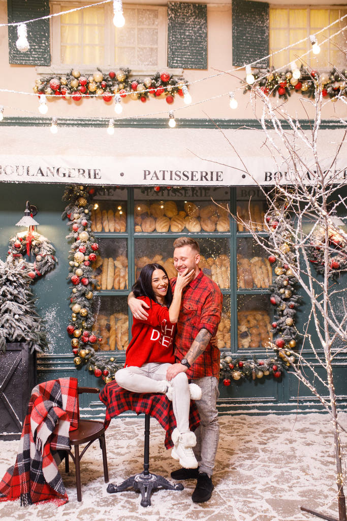 Ένα νεαρό όμορφο ζευγάρι, ένα μελαχρινό κορίτσι με κόκκινο πουλόβερ και λευκό παντελόνι, ένας άντρας με κόκκινο πουκάμισο κάθεται στους δρόμους του Παρισιού στο παρασκήνιο ενός παραθύρου ενός αρτοποιείου - Φωτογραφία, εικόνα