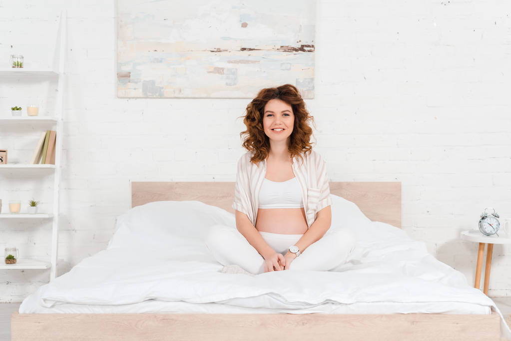 Piękna ciężarna kobieta uśmiecha się do kamery siedząc na łóżku  - Zdjęcie, obraz