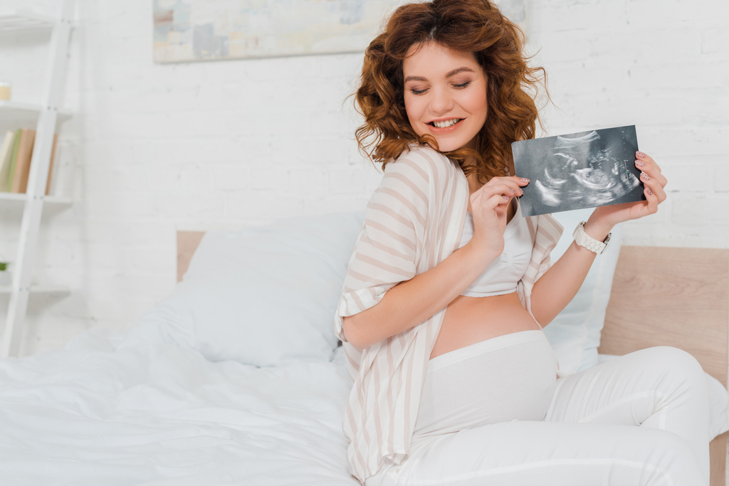 Atractiva mujer embarazada sonriendo mientras sostiene la ecografía del bebé en la cama
 - Foto, imagen